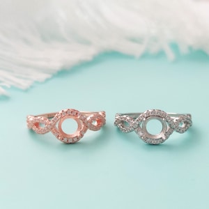 May include: Two engagement rings with a round center stone setting. The ring on the left is rose gold with a white stone setting. The ring on the right is silver with a white stone setting.