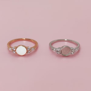 May include: Two rings with a round, white stone in the center. The ring on the left is gold with clear stones around the center stone. The ring on the right is silver with clear stones around the center stone.