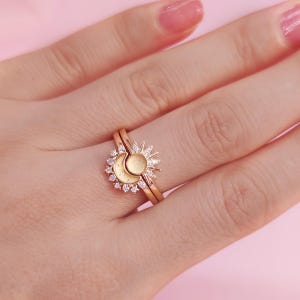 May include: A gold-colored ring set featuring a sun and moon design. The sun has a textured center and radiating rays, while the moon is a crescent shape. The rings are set with small, clear gemstones. The background is a soft pink.