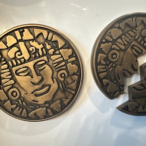 May include: Two gold-toned metal circular medallions with intricate designs. One medallion features a face with a headdress, while the other is broken into two pieces.