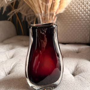 Könnte beinhalten: Eine kleine Glasvase mit schwerem Boden, die einen tiefen, gesättigten burgunderroten Kern aufweist, der von einer dicken Schicht aus klarem, transparentem Glas umschlossen ist. Die Vase hat eine glatte, abgerundete Form mit einem Hals.