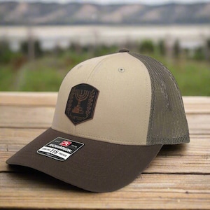 May include: A tan and brown trucker hat with a brown leather patch featuring a menorah and the text "State of Israel".