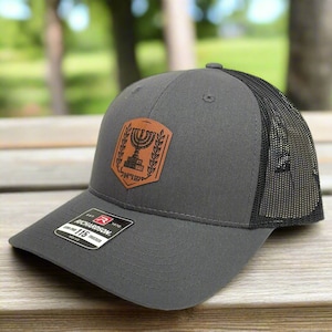 May include: A gray and black trucker hat with a brown leather patch featuring a menorah and the Hebrew word "Jerusalem".
