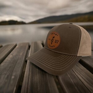 May include: An olive green baseball cap with a tan mesh back. The front features a circular brown leather patch with the text "EST. 1273 BCE" and "ISRAEL" surrounding a central graphic of the map of Israel with a Star of David symbol.
