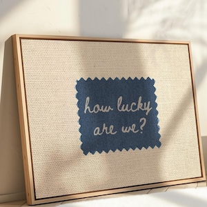 Pu&ograve; includere: Una stampa su tela incorniciata con sfondo in lino naturale. Una toppa di tessuto blu navy &egrave; centrata, con il testo bianco "how lucky are we?". La cornice &egrave; in legno chiaro e l'opera d'arte &egrave; appoggiata a un muro bianco.
