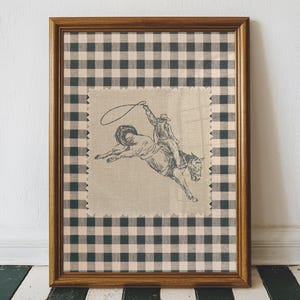 May include: A framed vintage-style artwork depicting a cowboy riding a bucking bronco. The image is set on a beige fabric patch, surrounded by a black and white gingham pattern, and enclosed in a wooden frame.