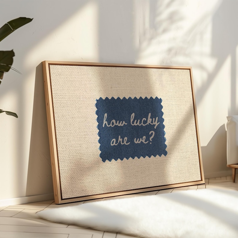 Pu&ograve; includere: Una stampa su tela incorniciata con sfondo in lino naturale. Una toppa di tessuto blu navy &egrave; centrata, con il testo bianco "how lucky are we?". La cornice &egrave; in legno chiaro e l'opera d'arte &egrave; appoggiata a un muro bianco.