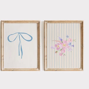 May include: Two framed art prints. One features a light blue bow on a white background. The other print has a floral arrangement in pink and purple hues on a striped background. Both are in gold-toned frames.