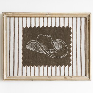 Puede incluir: Una obra de arte enmarcada con una ilustraci&oacute;n de un sombrero de vaquero. El sombrero est&aacute; dibujado en blanco sobre una tela marr&oacute;n oscuro, sobre un fondo de tela de rayas verticales. El marco es de color marr&oacute;n claro.