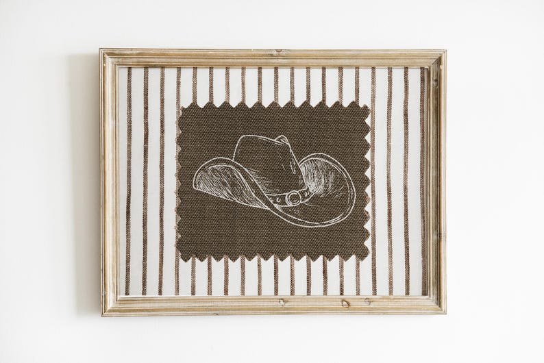 Puede incluir: Una obra de arte enmarcada con una ilustraci&oacute;n de un sombrero de vaquero. El sombrero est&aacute; dibujado en blanco sobre una tela marr&oacute;n oscuro, sobre un fondo de tela de rayas verticales. El marco es de color marr&oacute;n claro.