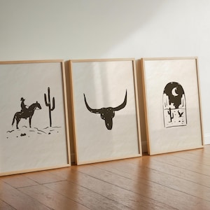 May include: Three framed art prints with a western theme. The prints feature a cowboy on a horse, a bull skull, and a desert landscape with a crescent moon. The artwork is in black and white.