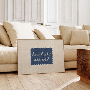Pu&ograve; includere: Una stampa su tela incorniciata con la scritta "how lucky are we?" su una toppa blu cucita. La stampa &egrave; ambientata in un soggiorno con un divano beige, cuscini ed elementi in legno. La combinazione di colori &egrave; prevalentemente neutra.