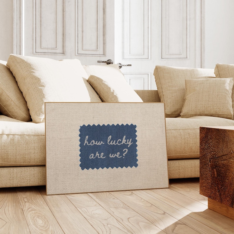 Pu&ograve; includere: Una stampa su tela incorniciata con la scritta "how lucky are we?" su una toppa blu cucita. La stampa &egrave; ambientata in un soggiorno con un divano beige, cuscini ed elementi in legno. La combinazione di colori &egrave; prevalentemente neutra.