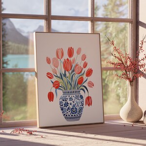 May include: A framed print of a watercolor painting of a bouquet of red and pink tulips in a blue and white patterned vase. The vase is sitting on a table in front of a window with a view of a lake and mountains.