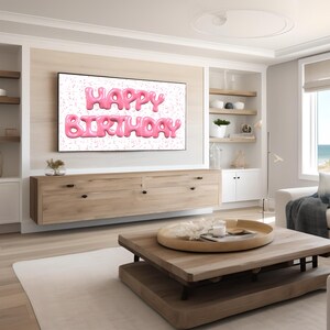 May include: A living room with a flat screen TV displaying the words "Happy Birthday" in pink balloon letters against a white background with pink confetti. The TV is mounted on a wall above a wooden cabinet. There is a wooden coffee table in front of a white couch with a blue throw blanket.