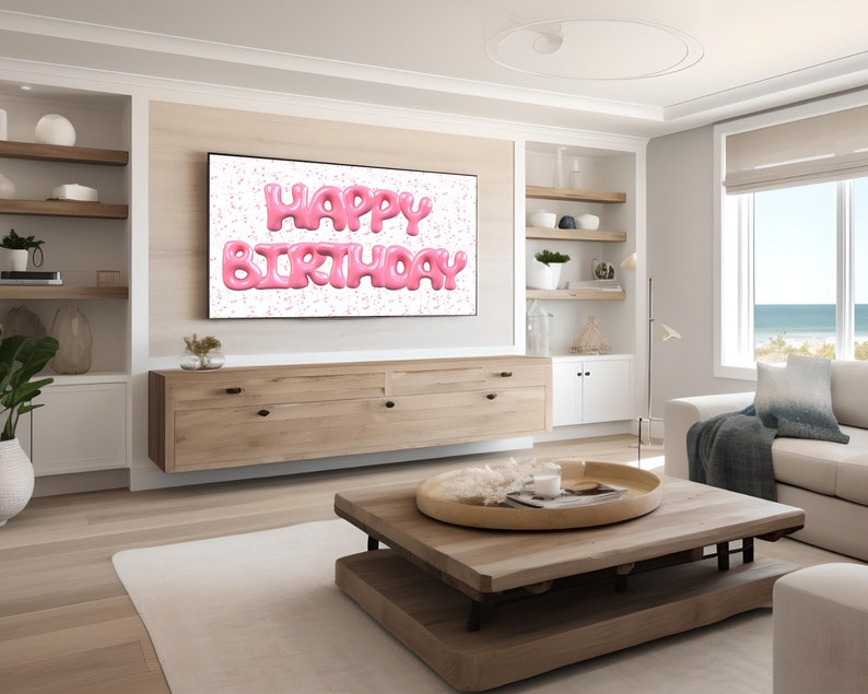 May include: A living room with a flat screen TV displaying the words "Happy Birthday" in pink balloon letters against a white background with pink confetti. The TV is mounted on a wall above a wooden cabinet. There is a wooden coffee table in front of a white couch with a blue throw blanket.