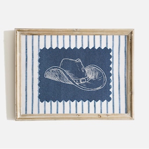 May include: A framed artwork featuring a blue and white striped background. A dark blue fabric patch with a white sketch of a cowboy hat is centered within the frame. The frame is a light gold color.