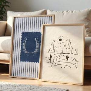 May include: Two framed art prints. One features a horseshoe design on a blue fabric patch over a striped background. The other print depicts a desert landscape with a road, cacti, and mountains, all in black and white.