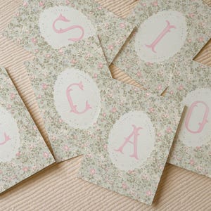 May include: A collection of rectangular cards featuring a light green background with a dense pattern of small pink roses and green leaves. Each card displays a large, pink, serif-style letter centered within an ornate, white, oval-shaped frame.