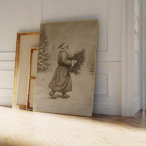 May include: A vintage black and white photograph of a man dressed as Father Christmas carrying a Christmas tree. The image is framed in a gold frame and is leaning against a white wall.