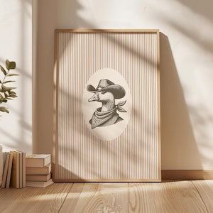 May include: Framed art print featuring a duck wearing a cowboy hat and bandana. The black and white illustration is set against a beige and white striped background, framed in natural wood. The artwork is displayed on a wooden floor.