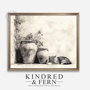 Op de afbeelding: Een ingelijste sepia-kleurige prent met een serene scène. Twee oude keramische vazen staan bij een muur met klimplanten. Een tabby kat ligt opgerold en slaapt vredig. De prent is ingelijst in een goudkleurige lijst. De tekst "KINDRED & FERN" staat onderaan.