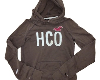 May include: A brown long-sleeve hooded sweatshirt featuring the white text HCO printed on the chest. Above the text is a small red embroidered bird logo. The garment includes a front kangaroo pocket and shows white paint-like marks on the sleeves.