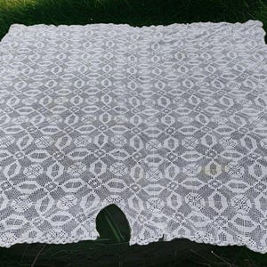 May include: White, open-weave lace tablecloth with a repeating geometric pattern. The edges are scalloped, and there is a small hole near the bottom. The tablecloth is displayed on a bed of green grass.