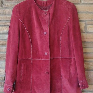 May include: A red suede jacket with a round collar and button closure. The jacket features white stitching along the seams and around the pockets. The sleeves have buttoned cuffs. The jacket is hanging on a wooden hanger.