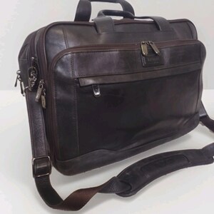 May include: A dark brown leather briefcase with a top handle and a detachable shoulder strap. The bag has multiple zippered compartments and silver-toned hardware. The leather appears worn, giving it a vintage look. The bag is designed for carrying documents and other essentials.