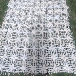 May include: A cream-colored, open-weave crochet blanket with a repeating geometric pattern. The blanket has a scalloped edge with fringe. The pattern features a series of octagonal shapes connected by smaller, star-like designs.