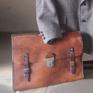 May include: A vintage brown leather briefcase with a metal clasp and two buckle straps. A gray tweed blazer is draped over the briefcase. The briefcase has a worn appearance, suggesting age and use.