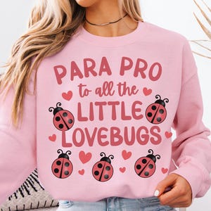 Pode incluir: Camisola rosa claro com o texto "PARA PRO to all the LITTLE LOVEBUGS" em vermelho e rosa. O design inclui joaninhas e corações. As palavras "Light Pink" estão no canto superior esquerdo.
