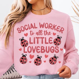 Pode incluir: Camisola rosa claro com o texto "SOCIAL WORKER to all the LITTLE LOVEBUGS" e gráficos de joaninhas. A camisola é de gola redonda e é feita de um material macio. O texto "Light Pink" está no canto superior esquerdo.