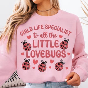 Pode incluir: Camisola rosa claro com o texto "CHILD LIFE SPECIALIST to all the LITTLE LOVEBUGS" e gráficos de joaninhas. A camisola é de cor rosa claro.