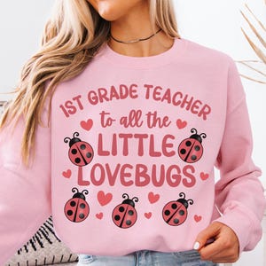 Pode incluir: Camisola rosa claro com o texto "1st Grade Teacher to all the LITTLE LOVEBUGS" e gráficos de joaninhas. A camisola é de estilo gola redonda e é feita de um material macio. O texto está em uma fonte lúdica.