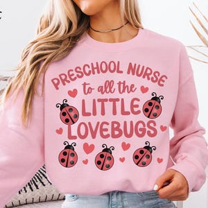 Pode incluir: Camisola rosa claro com o texto "Preschool Nurse to all the Little Lovebugs" em rosa. O design inclui joaninhas e corações. As palavras "Light Pink" estão no canto superior esquerdo.