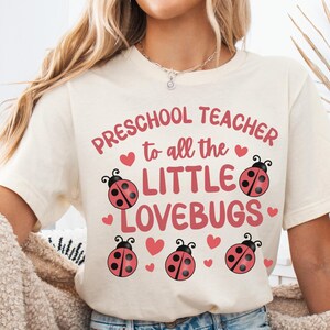 May include: Cream-colored t-shirt with the text "Preschool Teacher to all the LITTLE LOVEBUGS" in pink. The design includes pink hearts and ladybugs. The word "Natural" is printed in black in the upper left corner.