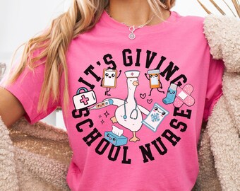 May include: A pink t-shirt with the text "IT'S GIVING SCHOOL NURSE" in a circular design. The design features a cartoon goose wearing a nurse's hat, surrounded by medical supplies. The words "Charity Pink" are in the upper left corner.