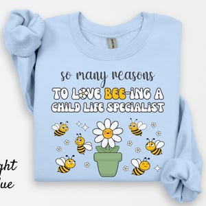 May include: Light blue sweatshirt with the text "so many reasons TO LOVE BEE-ING A CHILD LIFE SPECIALIST." The design includes cartoon bees, a daisy, and a flower pot.