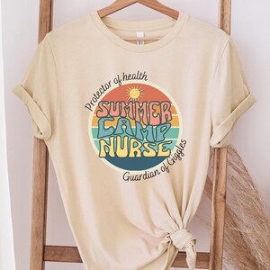 May include: A beige t-shirt with a retro-style graphic that says "Summer Camp Nurse" in a circle with a sun and rainbow stripes. The text around the circle says "Protector of health" and "Guardian of Giggles".