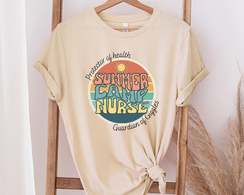 May include: A beige t-shirt with a retro-style graphic that says "Summer Camp Nurse" in a circle with a sun and rainbow stripes. The text around the circle says "Protector of health" and "Guardian of Giggles".