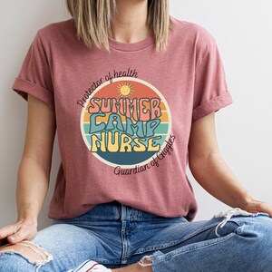 May include: A pink t-shirt with a retro-style graphic that says "Summer Camp Nurse" with a sun and rainbow design. The text below the graphic says "Protector of health, Guardian of Giggles".