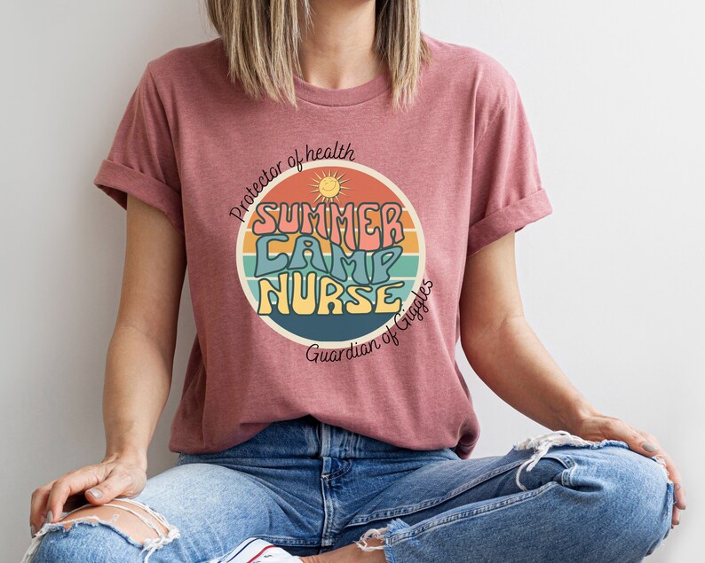 May include: A pink t-shirt with a retro-style graphic that says "Summer Camp Nurse" with a sun and rainbow design. The text below the graphic says "Protector of health, Guardian of Giggles".