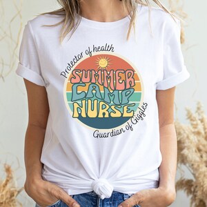 May include: White t-shirt with a retro-style graphic that says "Summer Camp Nurse" in colorful letters. The graphic is surrounded by a rainbow-colored circle with a sun and the text "Protector of health" and "Guardian of Giggles".
