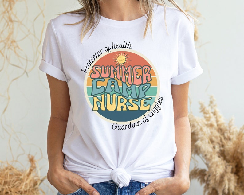 May include: White t-shirt with a retro-style graphic that says "Summer Camp Nurse" in colorful letters. The graphic is surrounded by a rainbow-colored circle with a sun and the text "Protector of health" and "Guardian of Giggles".