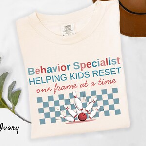 May include: Ivory-colored t-shirt with the words "Behavior Specialist HELPING KIDS RESET one frame at a time" in blue, red, and black. A bowling pin and ball graphic is below the text. The shirt is made of a soft material.