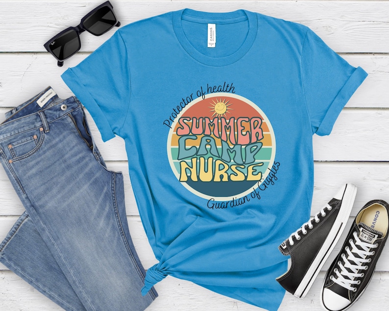 May include: A blue t-shirt with a retro-style graphic that says "Summer Camp Nurse" in colorful letters. The graphic also includes a sun and a rainbow. The text below the graphic says "Protector of health, Guardian of Giggles".