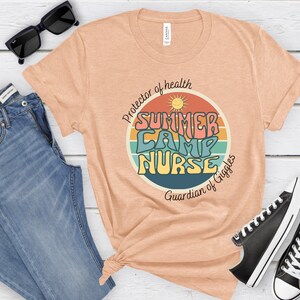 May include: A peach-colored t-shirt with a retro-style graphic that says "Summer Camp Nurse" in a colorful font. The graphic is surrounded by a rainbow-colored circle with a sun and the text "Protector of health" and "Guardian of Giggles".
