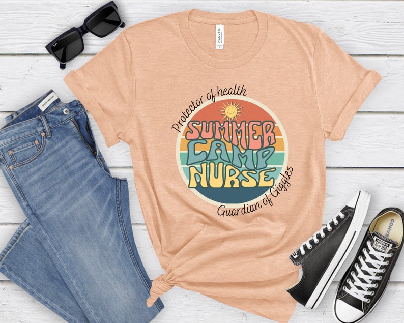 May include: A peach-colored t-shirt with a retro-style graphic that says "Summer Camp Nurse" in a colorful font. The graphic is surrounded by a rainbow-colored circle with a sun and the text "Protector of health" and "Guardian of Giggles".